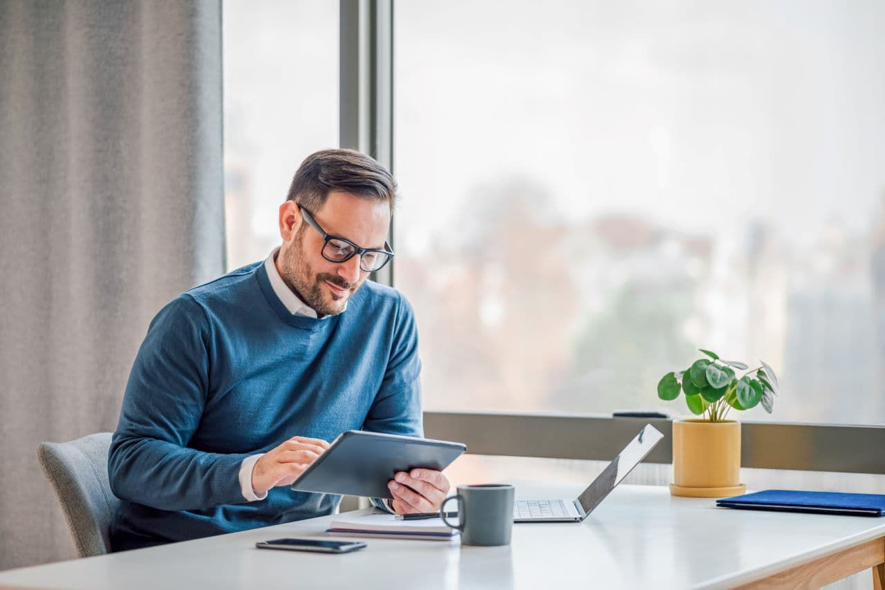Young businessman using digital tablet while working on laptop in business office. Male professional with wireless computer at desk. He is sitting by window in office.