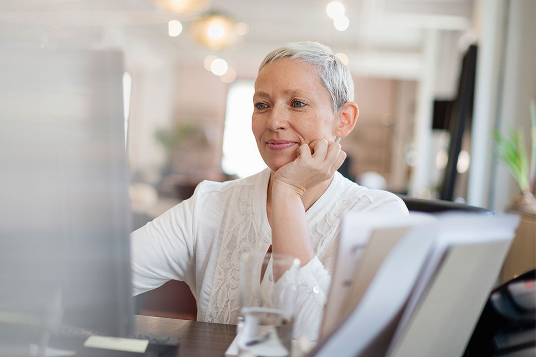 A woman smiling while her hand is folded against her face. She is looking at a computer screen. 