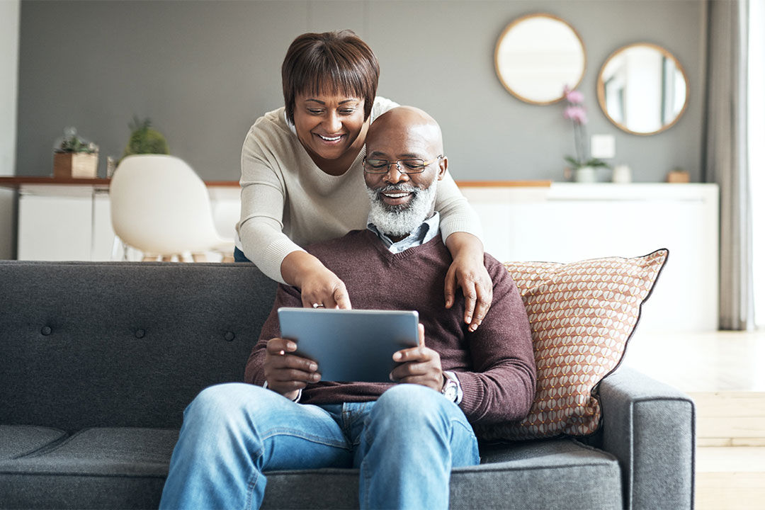 An older couple, a man and a woman, in a living room. They're both smiling. The man is sitting on the couch looking at his tablet. The woman is standing behind him, her arms are wrapped around him. She is also smiling and looking at the same tablet.  