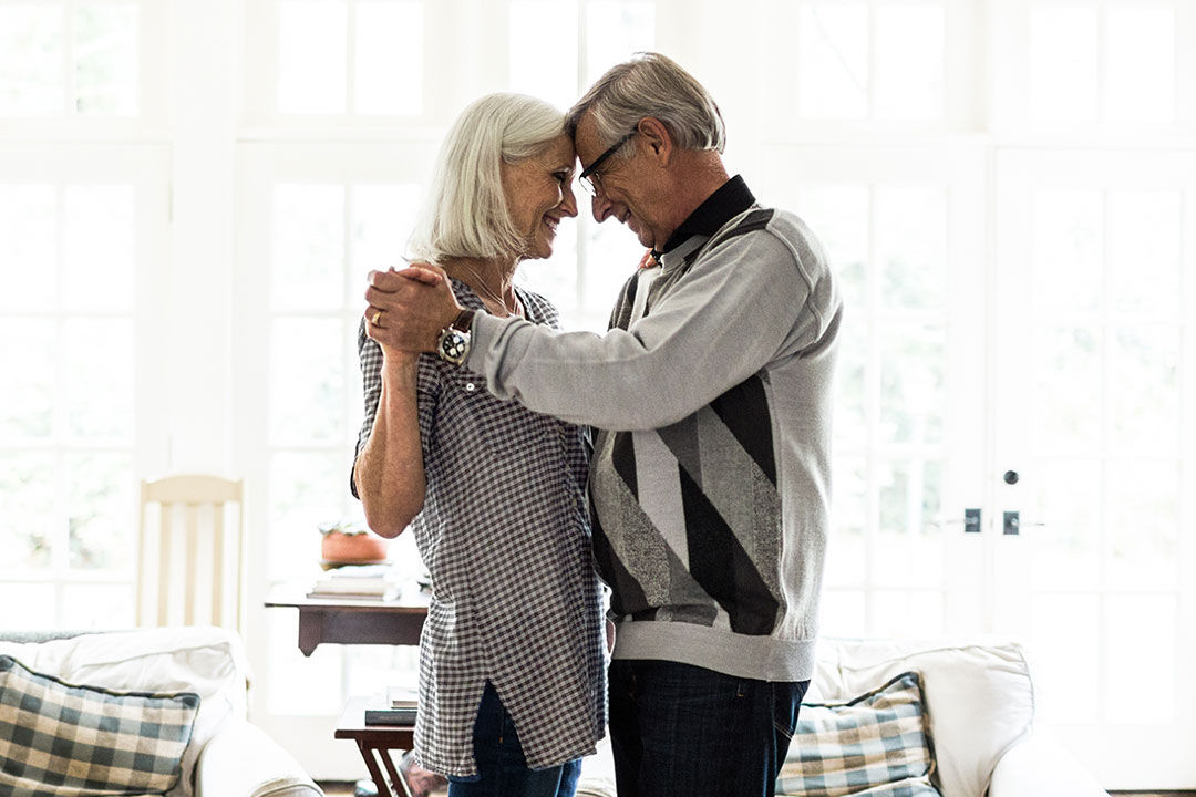 An older couple, a man and a woman, dancing in a living room against a large window. 