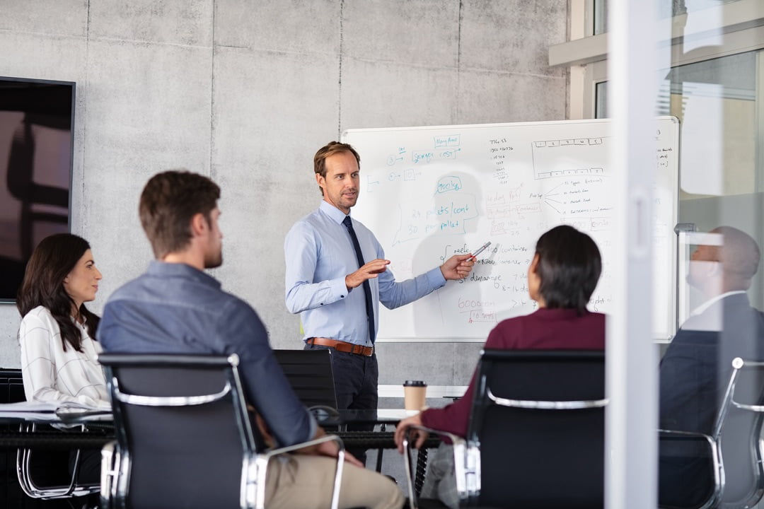 A man standing in front of a whiteboard. Four people are sitting and watching him speak.